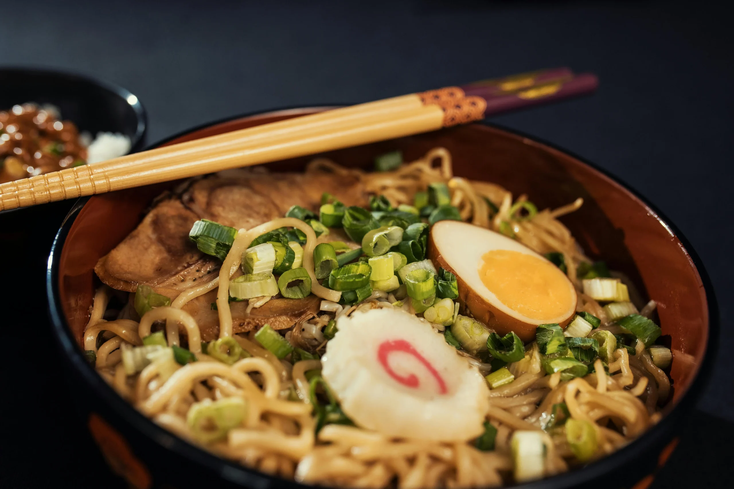 Bowl of Japanese ramen noodles with soft-boiled egg, green onions, and mushrooms, served with wooden chopsticks in a dark ceramic bowl