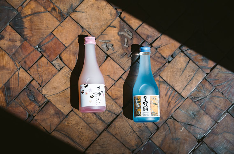 Two Japanese sake bottles on rustic wooden surface - a pink-labeled sake bottle and a blue-labeled sake bottle with traditional Japanese characters, showcasing different sake varieties for pairing-ele