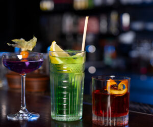 Three colorful drinks on a bar counter, including a purple cocktail in a coupe glass, a tall green mocktail with lemon and straw, and a red cocktail with an orange garnish.