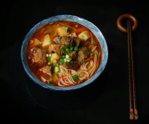 A bowl of ramen with noodles, tender beef chunks, tofu, and vegetables in a rich red broth, garnished with chopped green onions, served with wooden chopsticks.