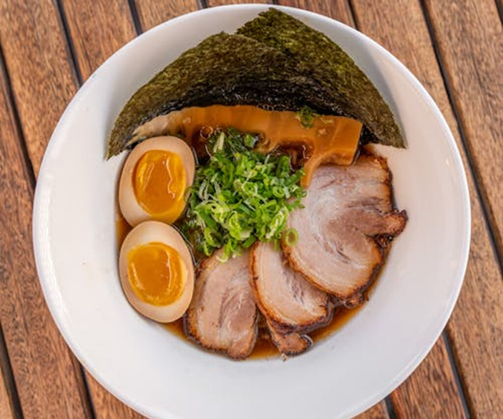 A bowl of Japanese ramen with sliced chashu pork, soft-boiled eggs, chopped green onions, bamboo shoots, and seaweed, served in a rich broth on a wooden table.