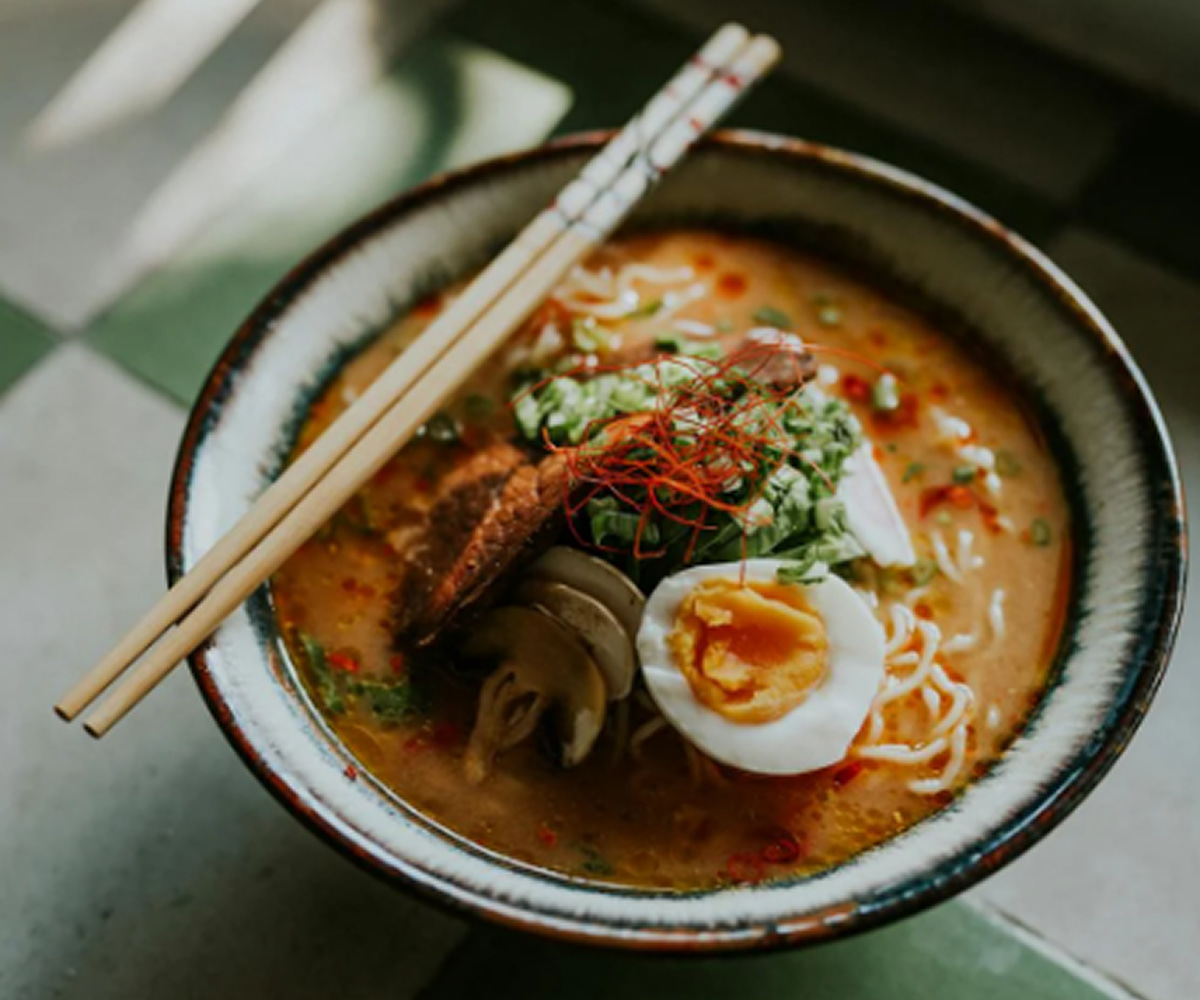 A bowl of ramen noodle soup with miso broth, fresh noodles, soft-boiled egg, grilled meat, mushrooms, and scallions, served with chopsticks.