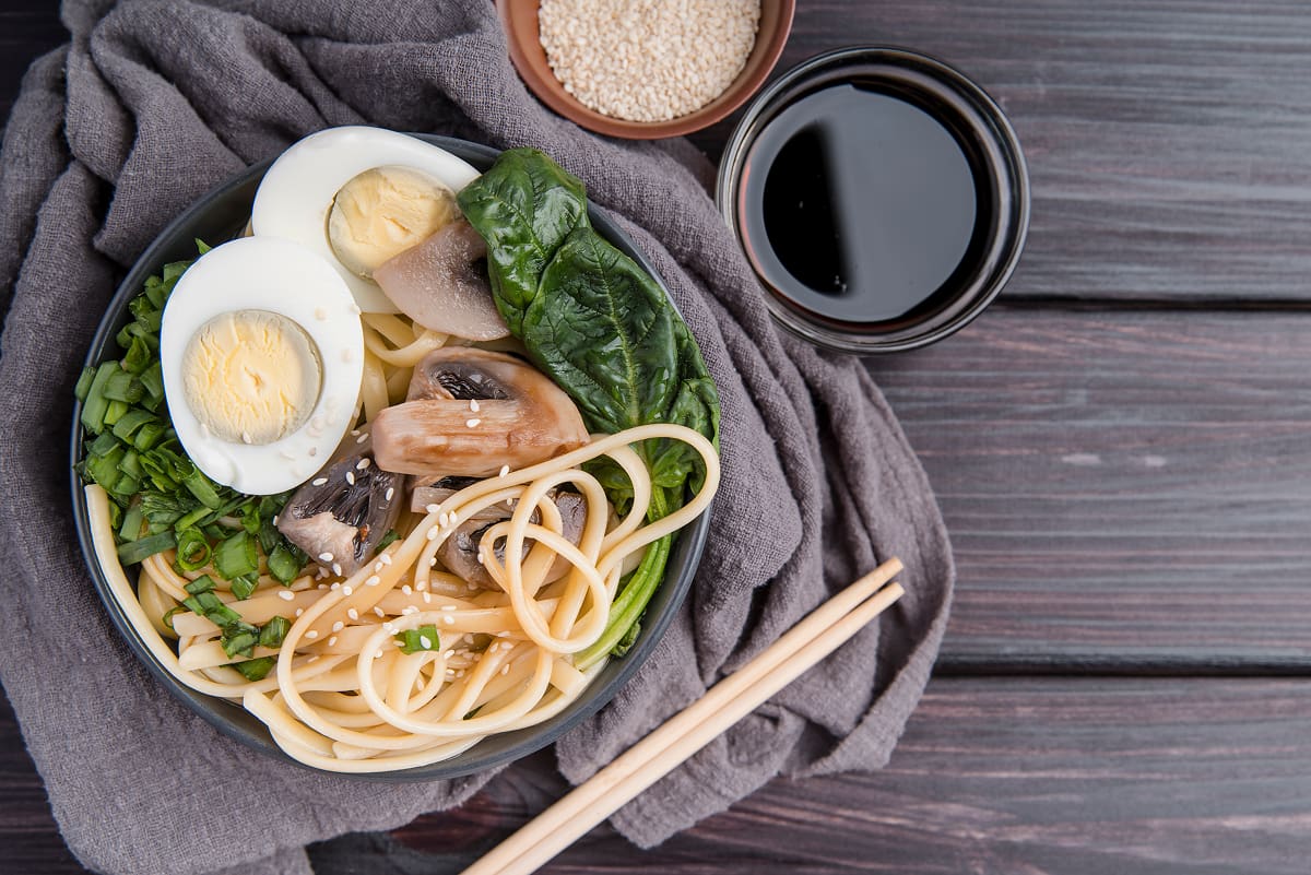 Ramen bowl with boiled eggs, spinach, mushrooms, noodles, and sesame seeds served with soy sauce and chopsticks on a wooden table.