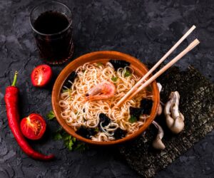 Japanese ramen bowl with shrimp, seaweed, and noodles served with chopsticks, fresh vegetables, and chili pepper on a dark textured background.