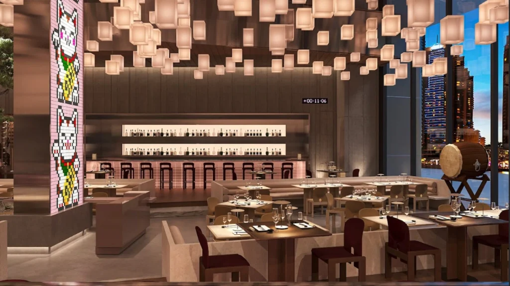 A modern, upscale restaurant interior featuring warm lighting and cube-shaped hanging lanterns. The space includes neatly set dining tables, a long illuminated bar with bottles displayed behind it, and large windows showing a nighttime city skyline. A tall column decorated with pixel-style lucky cat artwork stands on the left, and a large wooden barrel on a stand is visible on the right.