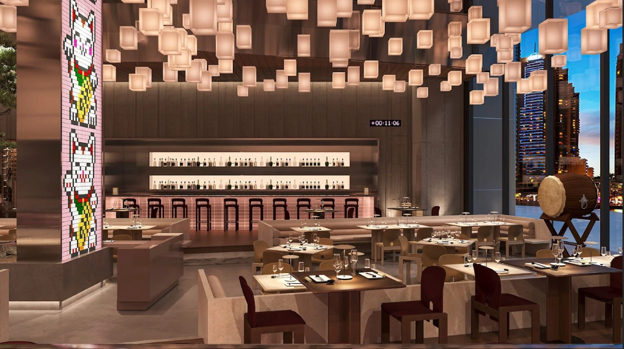 A modern, upscale restaurant interior featuring warm lighting and cube-shaped hanging lanterns. The space includes neatly set dining tables, a long illuminated bar with bottles displayed behind it, and large windows showing a nighttime city skyline. A tall column decorated with pixel-style lucky cat artwork stands on the left, and a large wooden barrel on a stand is visible on the right.
