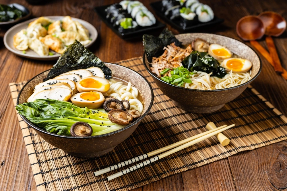 Bowls of Japanese ramen topped with soft-boiled eggs, sliced chicken, mushrooms, bok choy, seaweed, and green onions, served with sushi and gyoza on a wooden table at Ram & Roll, Dubai.