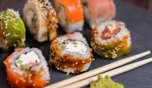 Close-up of assorted sushi rolls topped with sesame seeds, colorful fish, and avocado, arranged on a dark slate plate. A pair of wooden chopsticks and a small portion of wasabi are placed beside the rolls.
