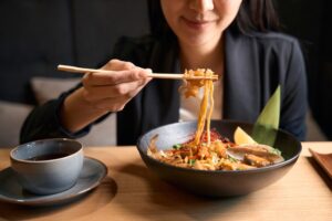 A woman uses chopsticks to lift noodles from a bowl of ramen, with toppings like vegetables and mushrooms, while sitting at a wooden table with a cup of tea beside her.