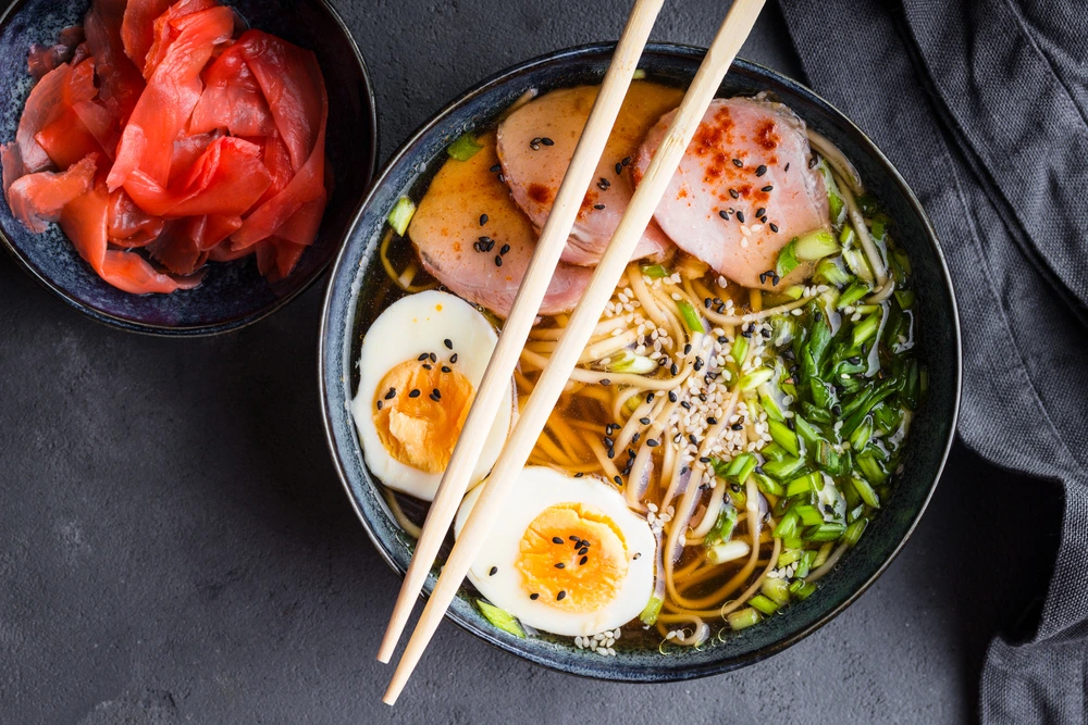 Japanese ramen bowl with sliced chicken, soft-boiled egg, noodles, and fresh greens served with chopsticks